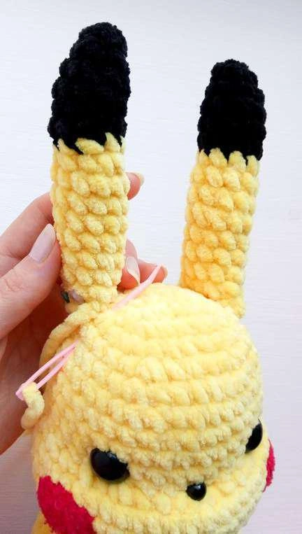 Hands pinning yellow crocheted ears with black tips to the top of a yellow Pikachu amigurumi head using pink sewing pins.