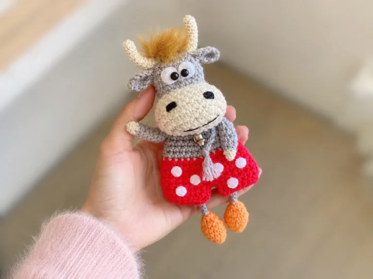 Hand holding a gray crochet bull with fluffy hair and red polka dot shorts using playful bull amigurumi pattern free.