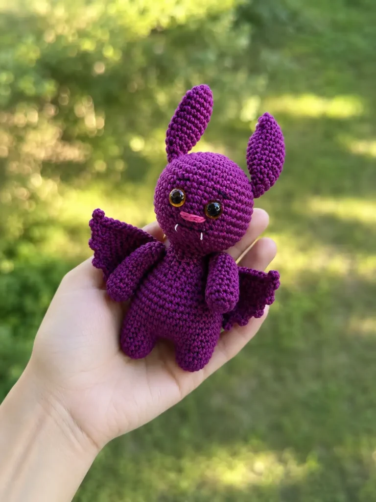 Crocheted purple Halloween bat plushie held in a hand featuring scalloped wings, amber safety eyes, tiny white fangs, and a pink embroidered nose.