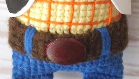 Close-up of a crocheted Sheriff Woody doll's midsection, showing a thick brown belt with a shiny oval buckle, blue embroidered belt loops, blue jeans, and a yellow shirt with orange plaid stitching.