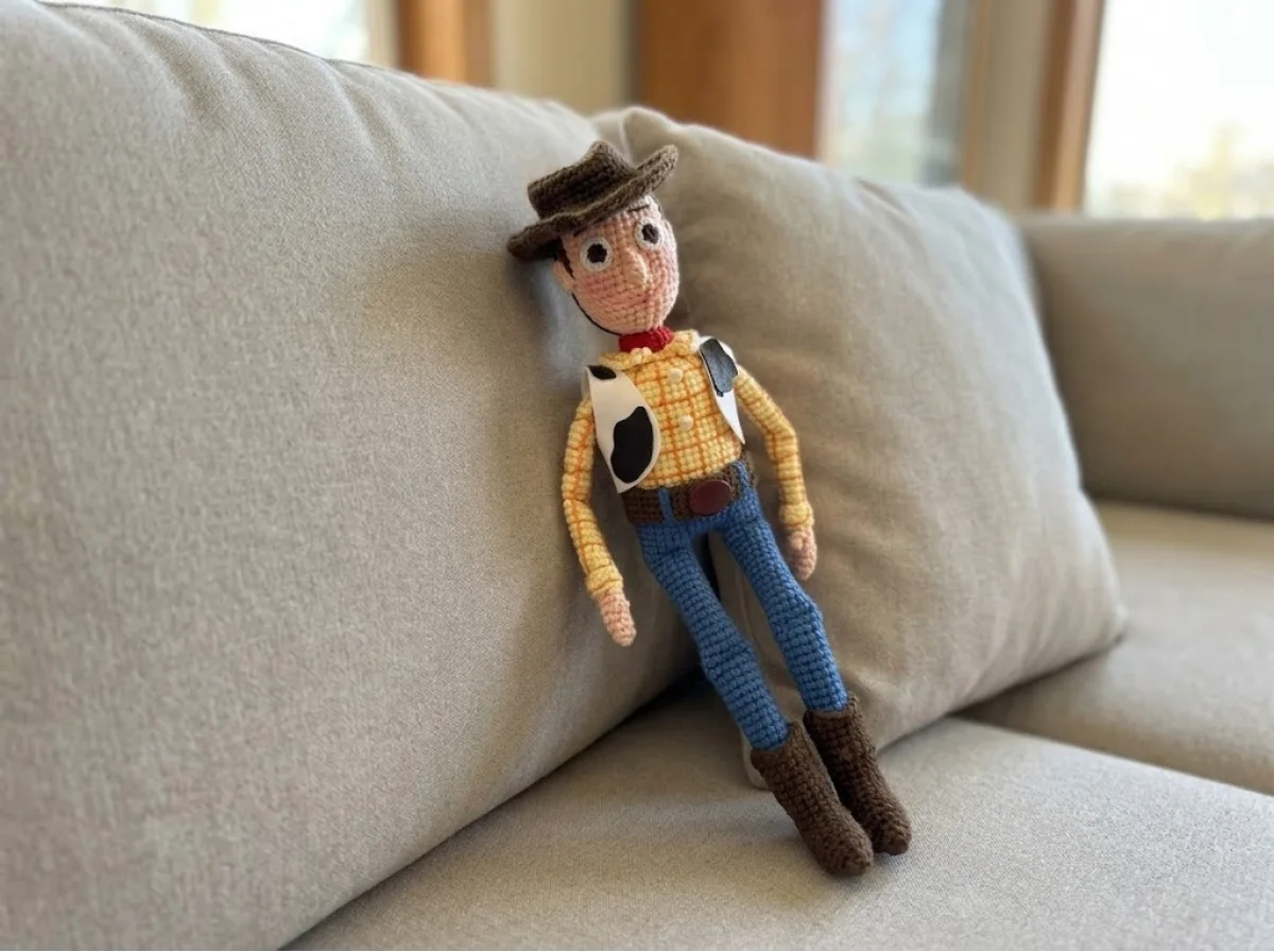 Crochet sheriff woody amigurumi free pattern sitting against a beige pillow, showing yellow shirt, cow vest, and brown hat.