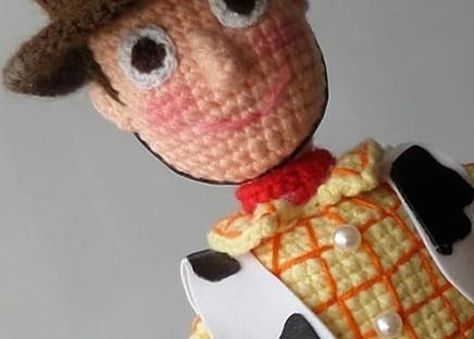 Close-up of a crocheted Sheriff Woody amigurumi doll showing a peachy face, brown hat, round white eyes, red neckerchief, and yellow shirt with orange plaid embroidery.