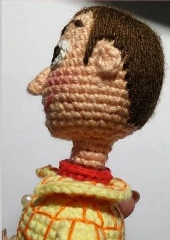 Side profile of a crocheted Sheriff Woody amigurumi doll without his hat, showing his brown yarn hair, attached crocheted ear, prominent nose, red neckerchief, and yellow plaid shirt.