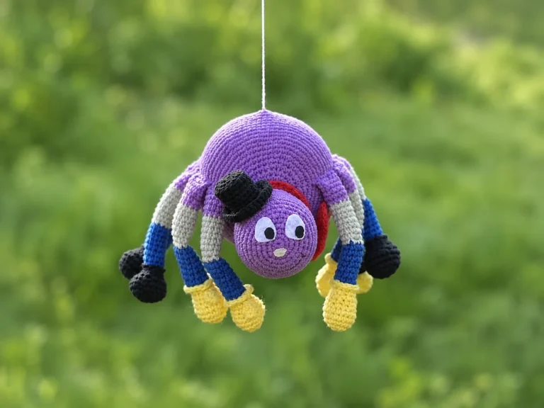 A crochet spider amigurumi wearing a black top hat and red bow tie, with color-blocked legs in purple, blue, and gray.