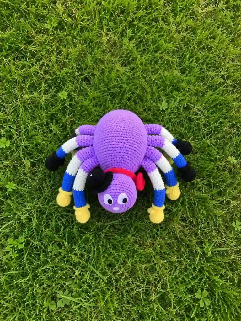 Top-down aerial view of a finished purple spider Porfiriyich amigurumi on green grass, displaying all eight color-blocked legs spread out.