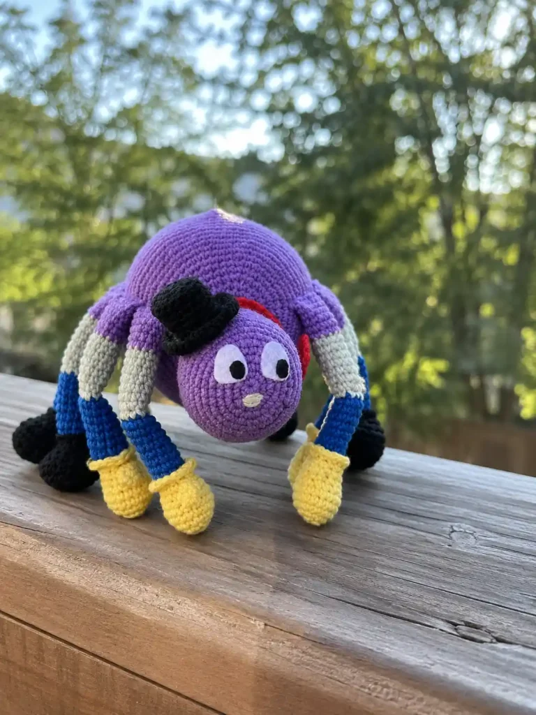 Handmade purple crochet spider Porfiriyich toy perched on a weathered wooden porch railing with a blurred green outdoor background.