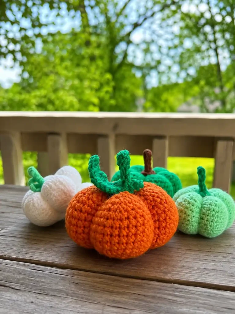 Four handmade crochet pumpkins in white, orange, dark green, and light green resting on a wooden deck surface, with a softly blurred background of green trees and a wooden railing.