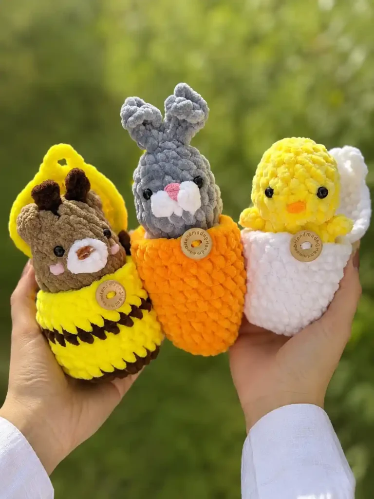 Two hands holding a set of three mini crochet animals tucked inside squishy yarn eggs against a blurred green background, featuring a bear in a bee egg, a bunny in a carrot egg, and a chick in a white egg.