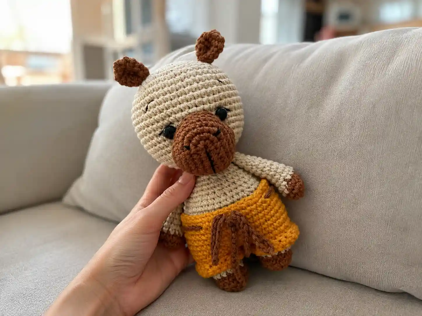 A beige and brown crochet capybara amigurumi wearing mustard yellow shorts, held in a hand indoors.