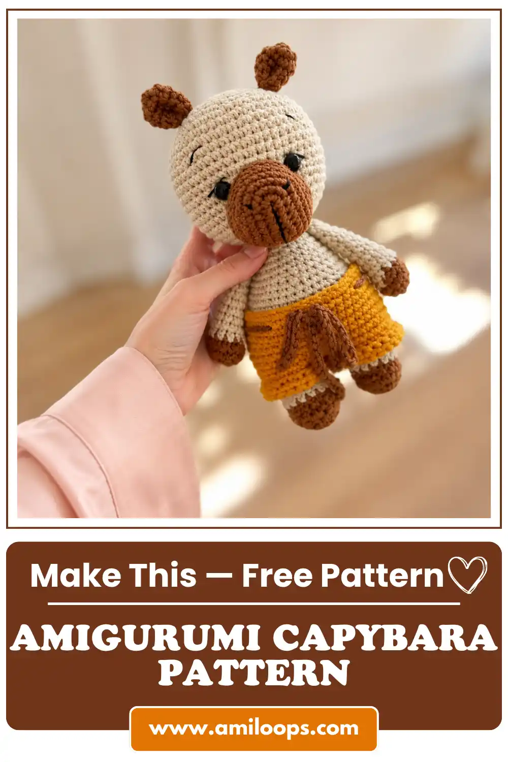 Hand in a pink sleeve holding an amigurumi capybara pattern toy with mustard shorts in a bright room.