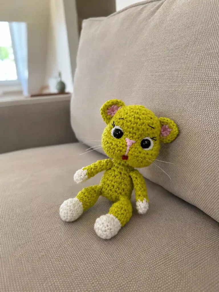 Completed avocado cat amigurumi pattern sitting upright on a beige couch cushion in a bright living room.