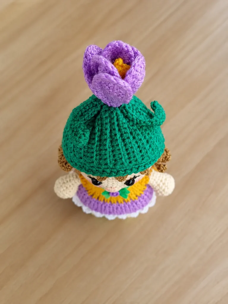 High-angle, top-down view of the baby crocus amigurumi doll resting on a wooden surface, highlighting the details of the green ribbed hat, tiny green leaves, and the vibrant purple crocheted crocus flower with yellow stamen on top.