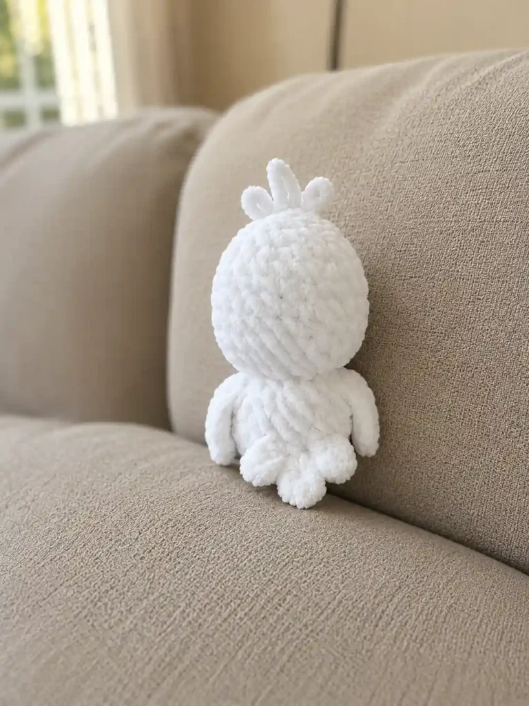 Back view of a white yarn baby goose amigurumi pattern toy showing its tail and fluffy head tuft on a beige sofa.