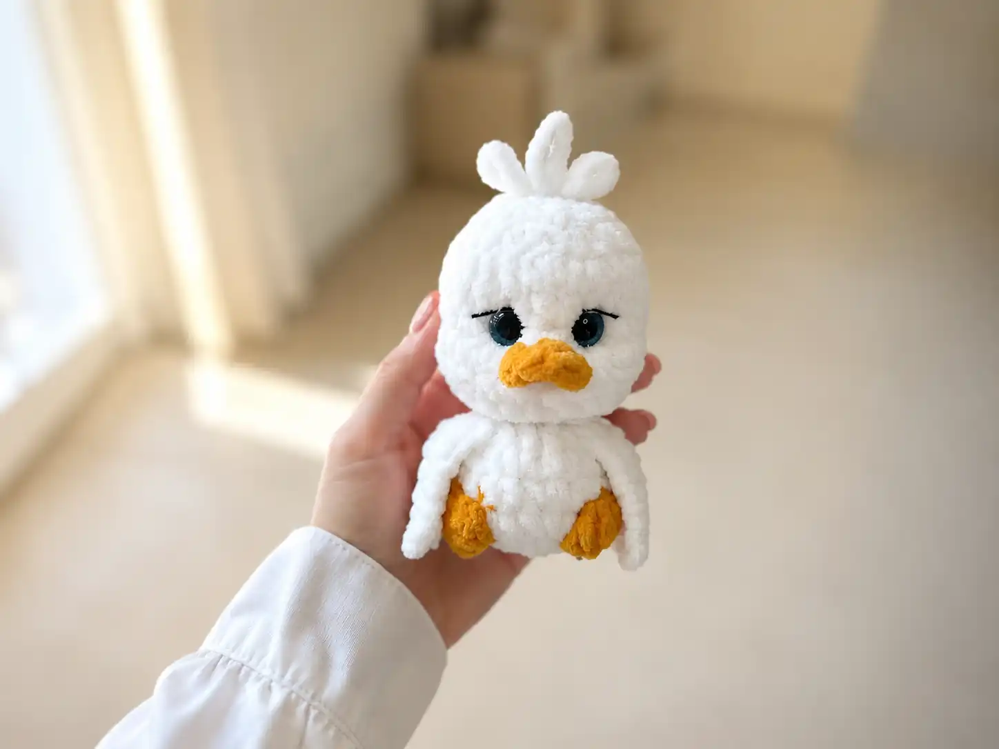 A small white crochet goose amigurumi with an orange beak and feet sitting on a beige couch cushion.