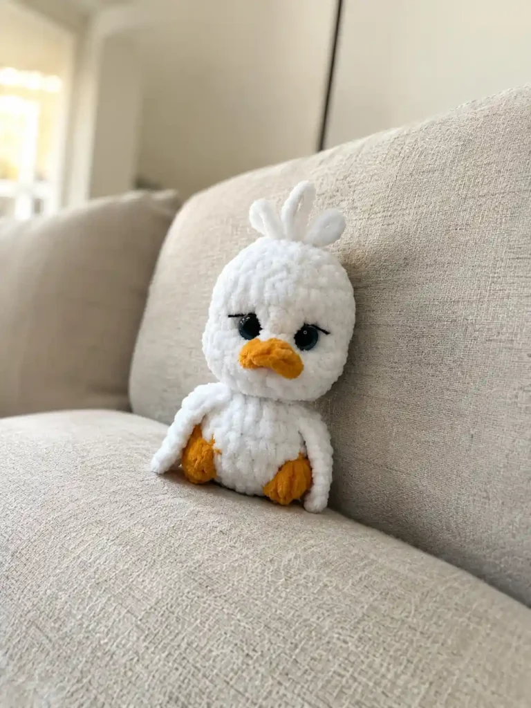 Front view of a soft white plush baby goose amigurumi pattern toy with orange beak and feet sitting on a beige sofa.