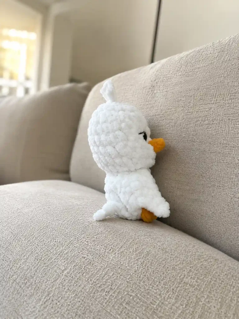 Side profile of a fluffy white baby goose amigurumi pattern toy featuring an orange beak resting on a beige sofa.