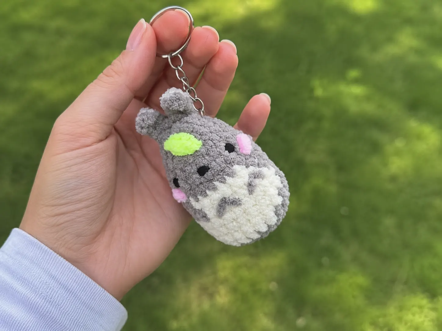 A gray and cream crochetTotoro Amigurumi Keychain with pink cheeks and a green leaf accent, held in a hand.