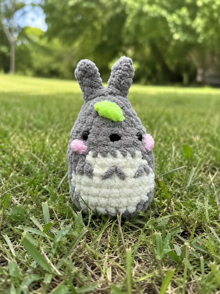 Fluffy gray yarn baby totoro crochet keychain free pattern sitting in green grass with pink cheeks and a green leaf.