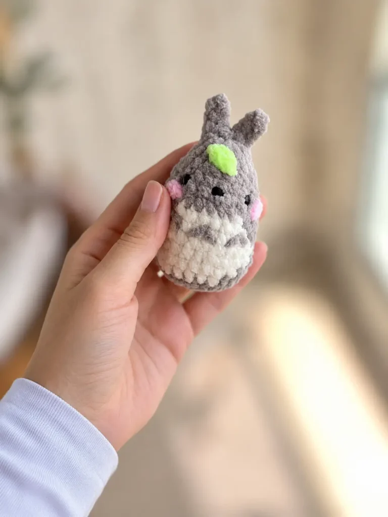 A hand holding a small gray baby totoro crochet keychain free pattern indoors with bright sunlight in the background.