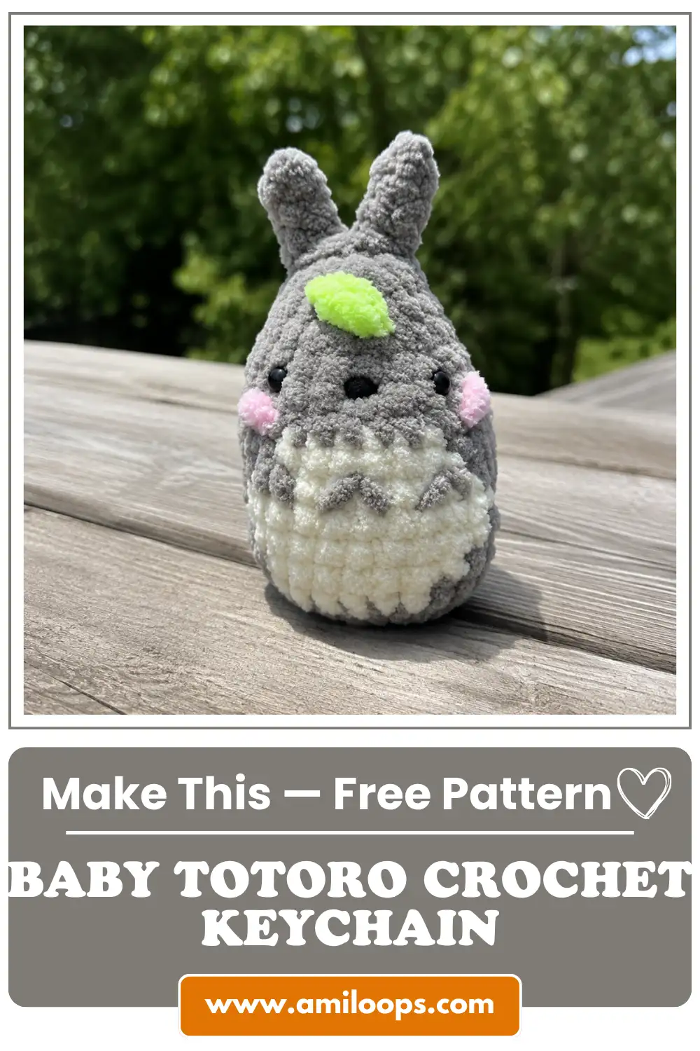 Grey and white yarn baby totoro crochet keychain free pattern sitting on a wooden table with blurred green trees behind it.
