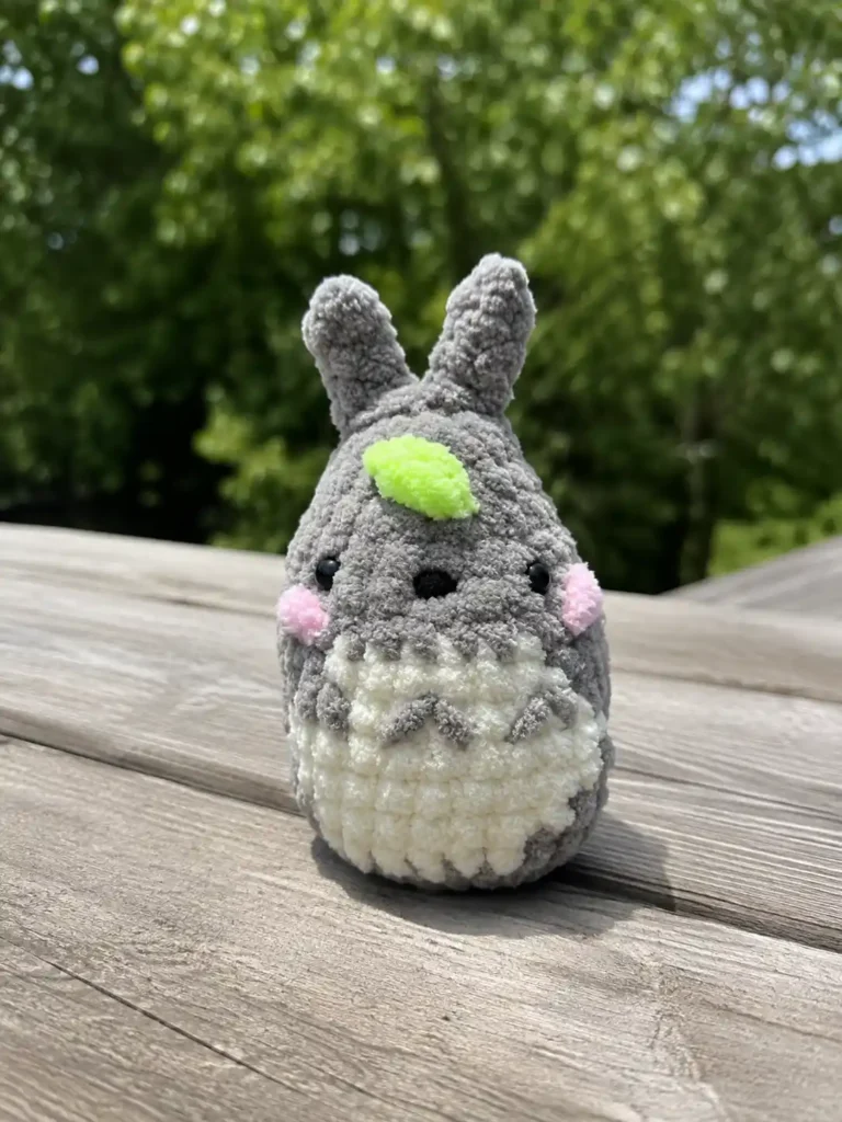 Finished baby totoro crochet keychain free pattern made of gray chenille yarn sitting on a wooden deck with trees behind.