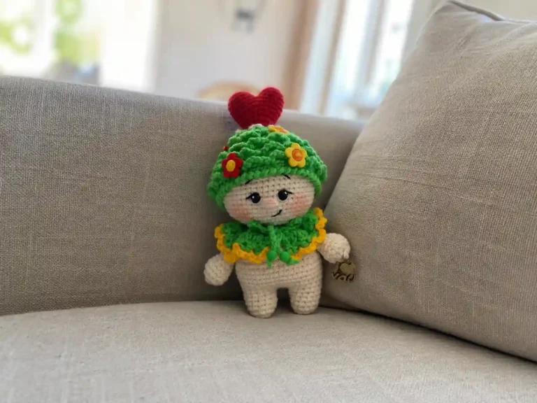 A crocheted baby doll wearing a textured white hat and beaded collar, holding a red crochet heart.