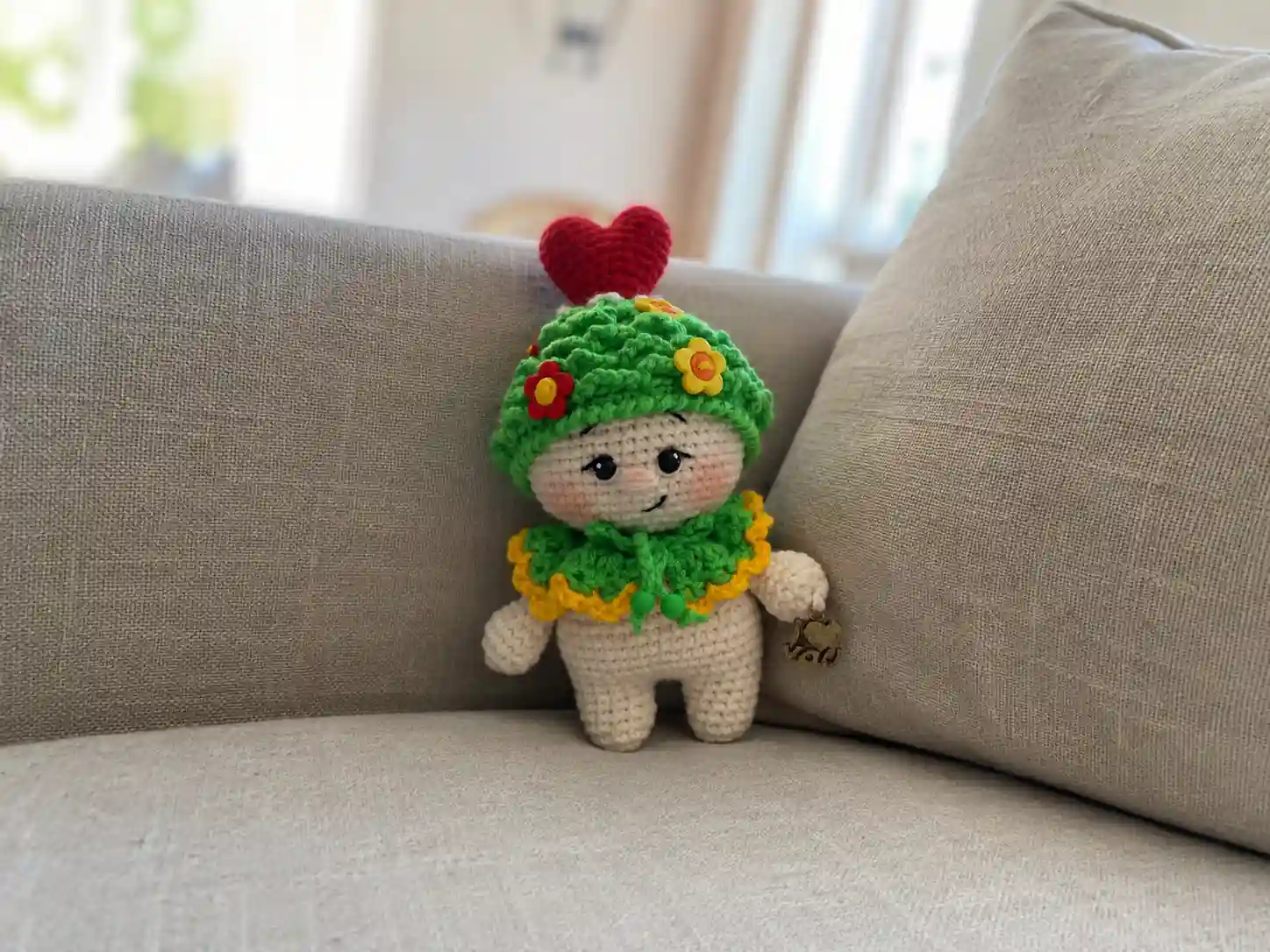 A crocheted baby doll wearing a textured white hat and beaded collar, holding a red crochet heart.