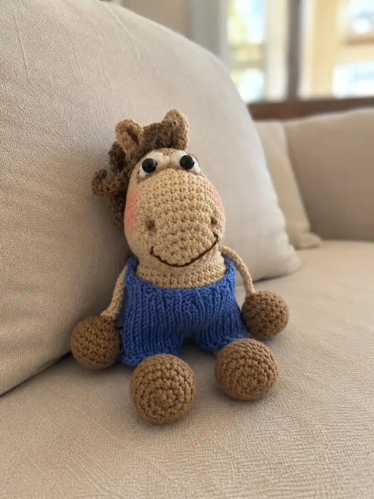 Caramel yarn toy from our amigurumi horse free pattern wearing blue knit pants, sitting on a light beige couch.