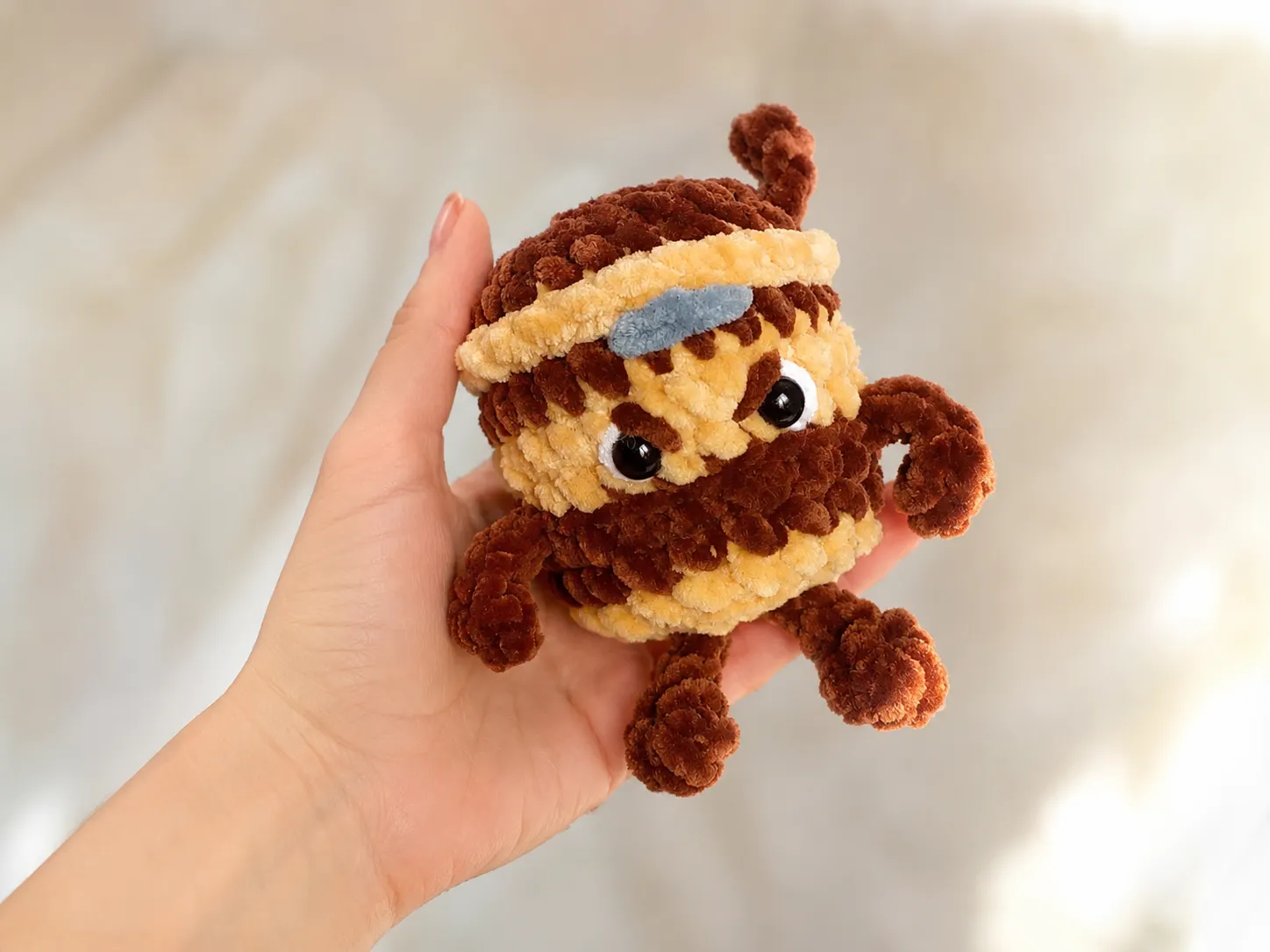 A hand holding a brown and beige crochet Cappuccino Assassino keychain with a gray felt headband.
