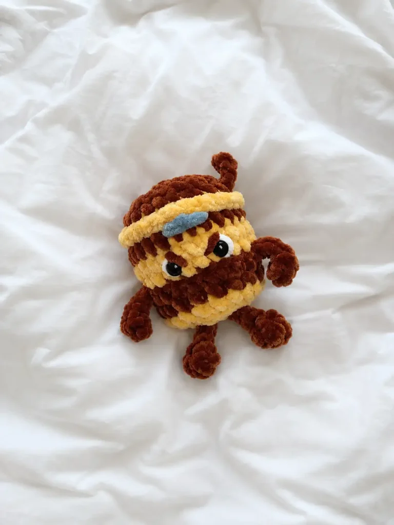 Handmade brown and beige chenille toy from the cappuccino assassino crochet pattern free lying on white bed sheets.