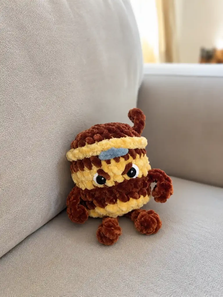 Amigurumi brown and beige ninja cup made using this cappuccino assassino crochet pattern free, resting on a grey sofa.