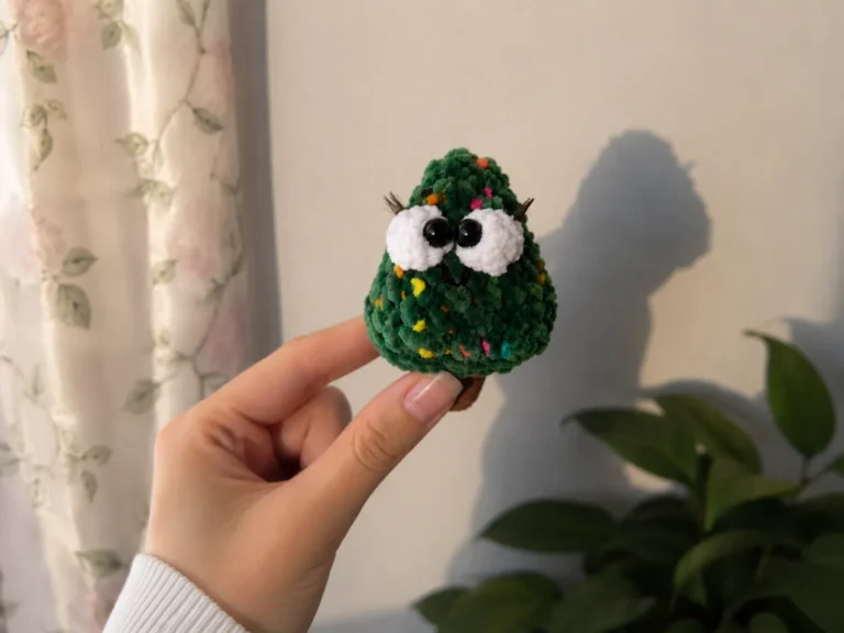 A hand holding a mini crochet Christmas tree amigurumi with white eyes and green chenille yarn.
