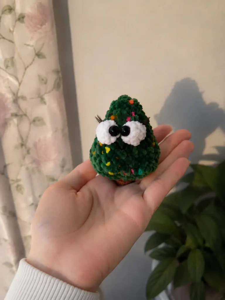 Hand holding a tiny green christmas tree amigurumi with a brown trunk, large white eyes, and lashes indoors near a plant.