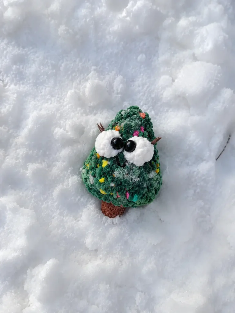 Green fluffy christmas tree amigurumi with big eyes and a brown trunk sitting in bright white outdoor snow.