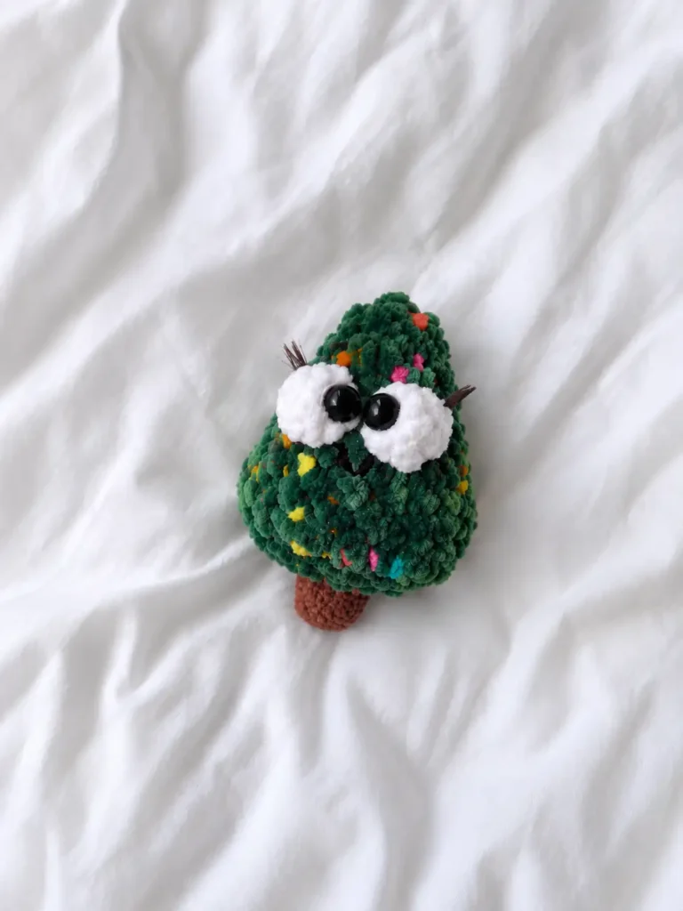 Small green fluffy christmas tree amigurumi with big white puff eyes and eyelashes resting on a white wrinkled bedsheet.