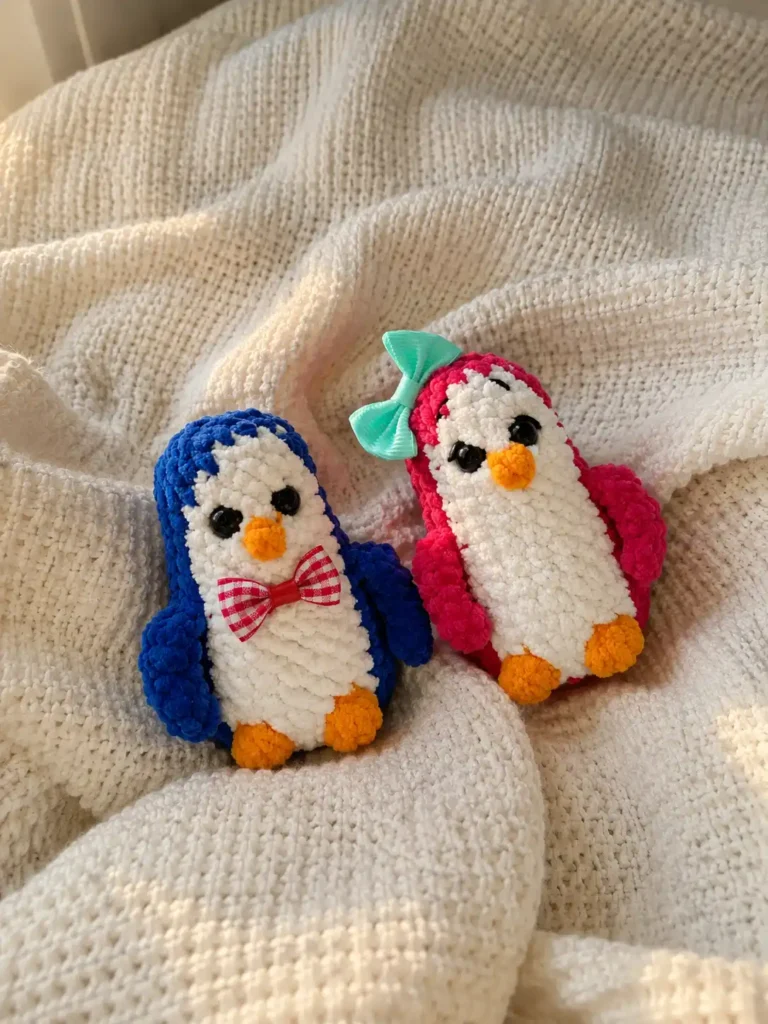 A blue and a pink amigurumi penguin made of thick chenille yarn resting on a textured white blanket.