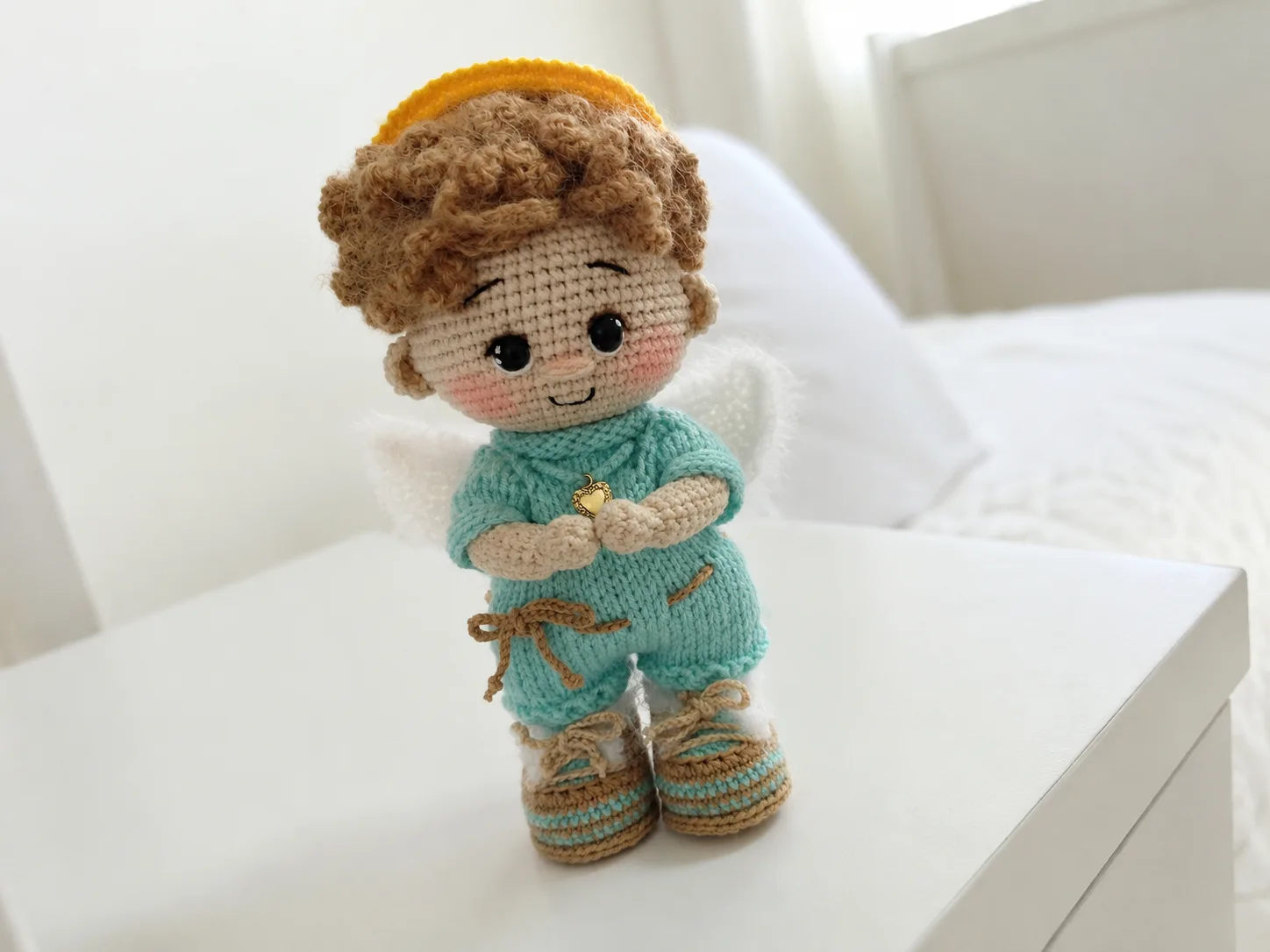A small crochet angel baby doll amigurumi with white wings, golden halo, and curly wig, held in a hand.