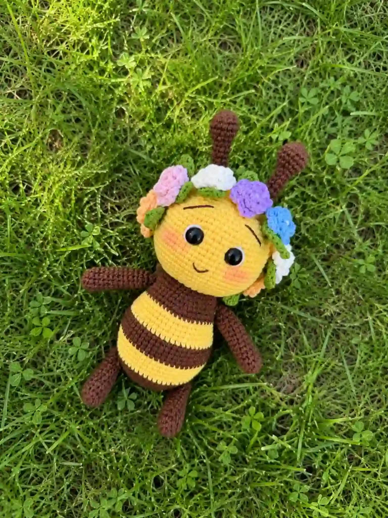Cute crochet bee pattern plush toy with brown yarn arms and flower crown lying outdoors on green grass.