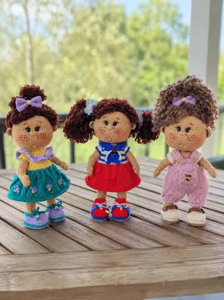 Three dolls in teal, red, and pink on an outdoor slatted table modeling a crochet boucle yarn wig free pattern.
