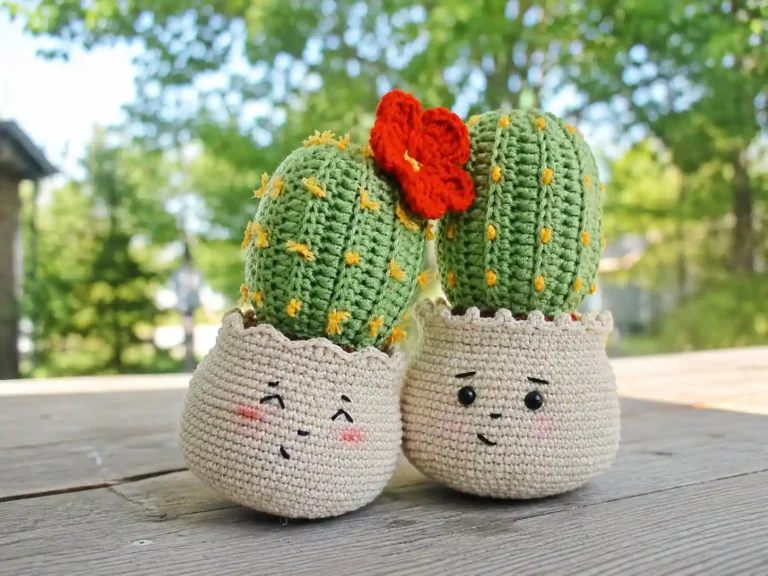 Two crochet cactus amigurumi in beige pots with embroidered faces, one topped with a red crochet flower, on a windowsill.