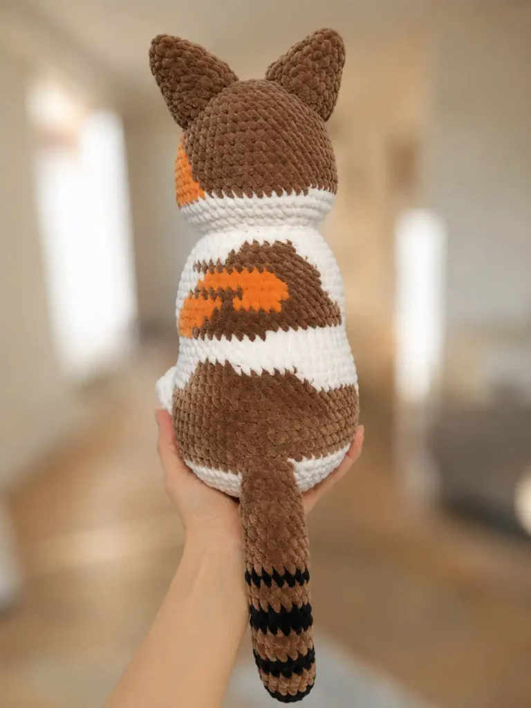 Back view of the crochet cat amigurumi free pattern in brown and white chenille yarn, showing a striped tail and patches.
