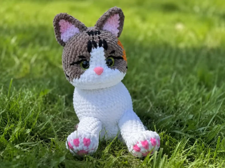 A pink and cream crochet cat amigurumi with textured bobble-stitch paws sitting upright.