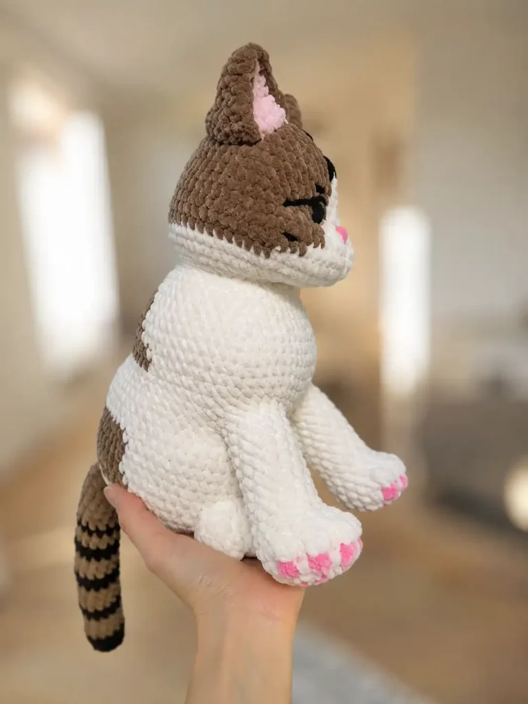 Side profile of a fluffy crochet cat amigurumi free pattern held in hand, highlighting pink toes and brown back stripes.
