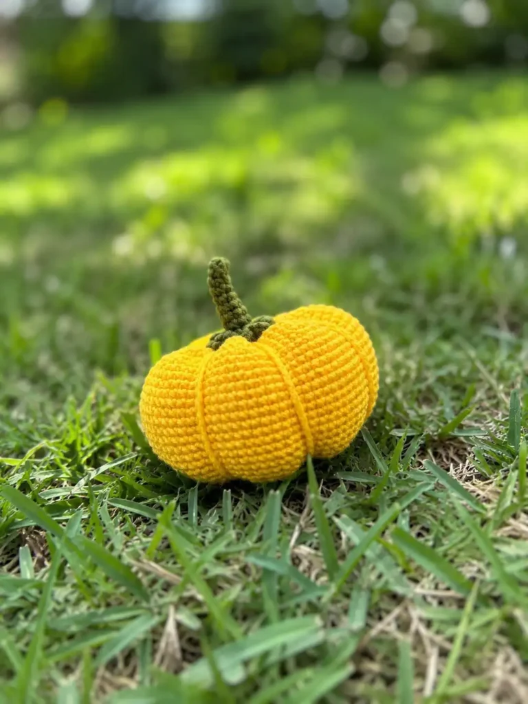 A yellow amigurumi made using this crochet pumpkin free pattern sitting directly on green grass in the sunlight.