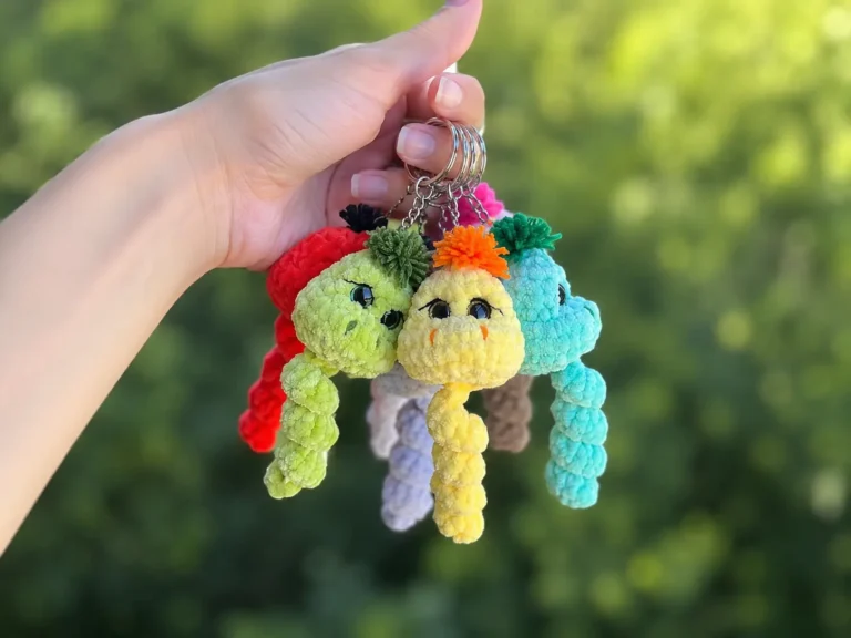 A hand holding several colorful crochet snake keychains on a keyring outdoors, with more shown in grass and on wood.