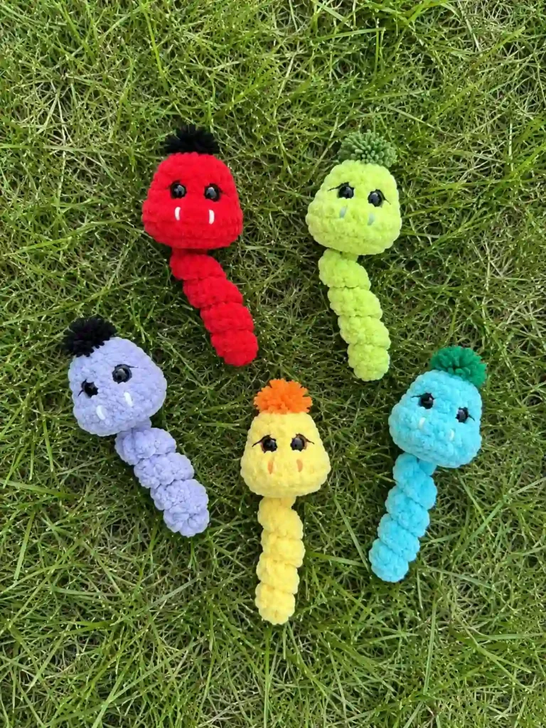 Five plush yarn snakes (red, green, purple, yellow, blue) on grass. A colorful crochet snake keychain pattern display.