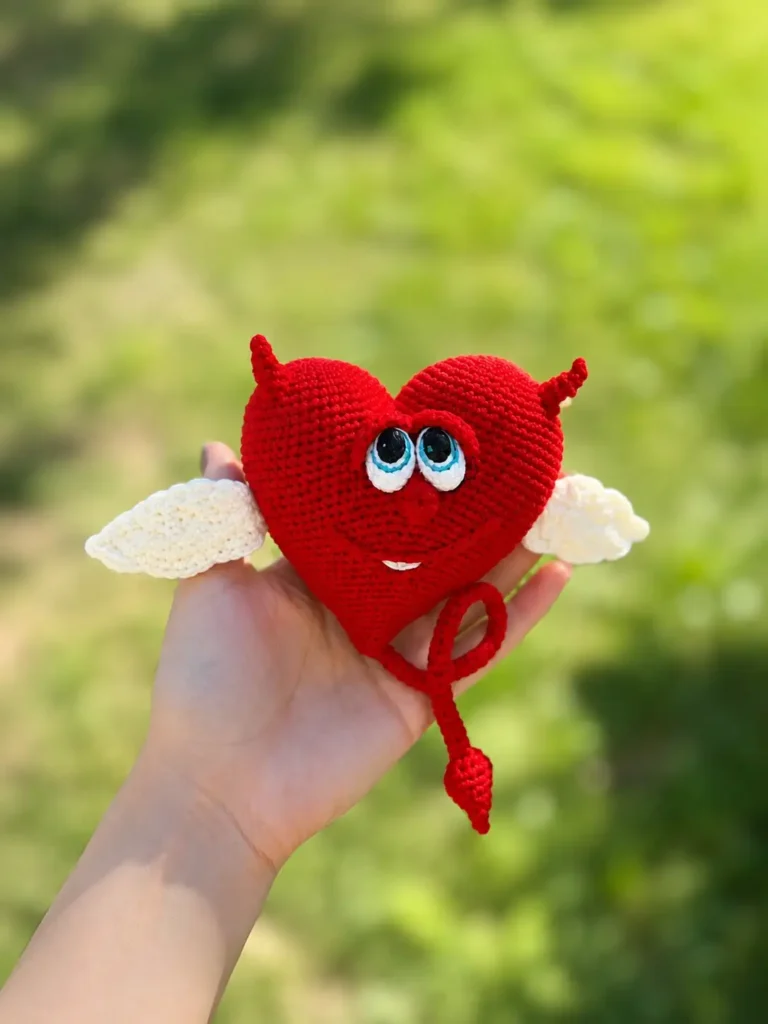 Hand holding a red yarn toy from our crochet valentine heart free pattern against a blurred green nature backdrop.