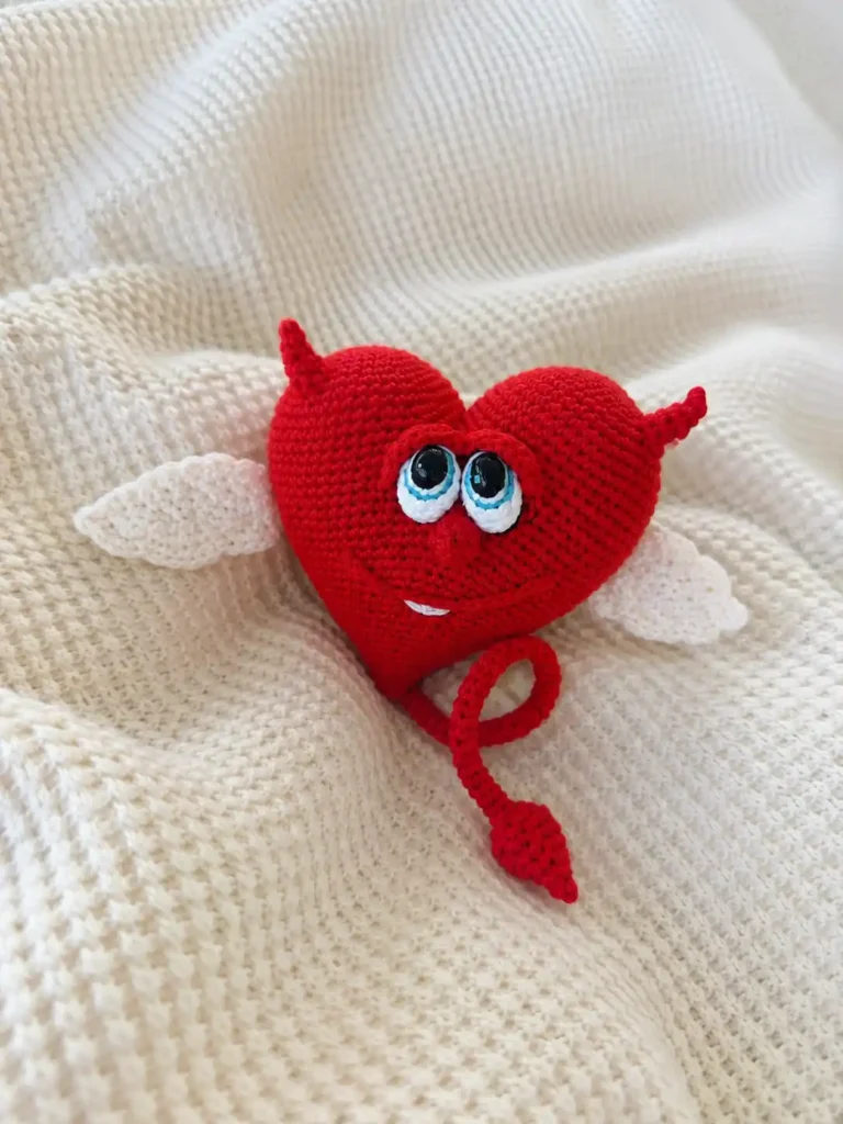Red yarn toy from our crochet valentine heart free pattern with white wings and a tail, sitting on a beige sofa.