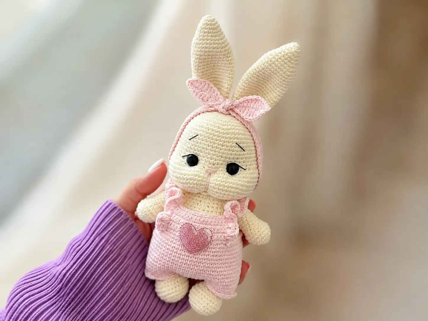 Cream crochet bunny amigurumi in pink overalls and ear bow, held against a beige couch.