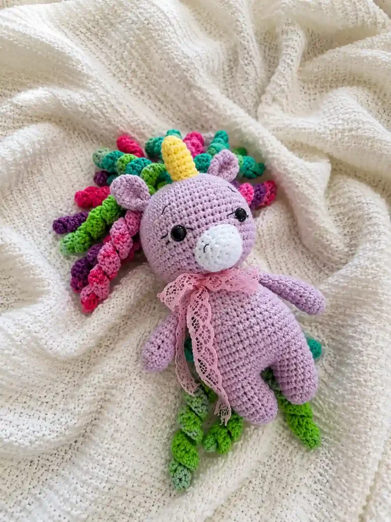 A purple cute unicorn amigurumi free pattern toy with a pastel curled mane resting softly on a white textured knit blanket.
